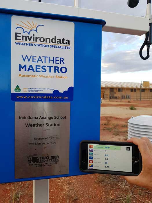 Phone displaying live readings such as temperature, humidity, wind speed, and other metrics next to the Indulkana School Weather Station.