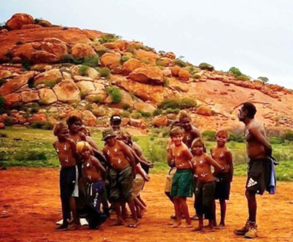 Gathered outdoors on red earth, with striking ochre rock formations rising behind them, the colours of the land, and the sense of community.